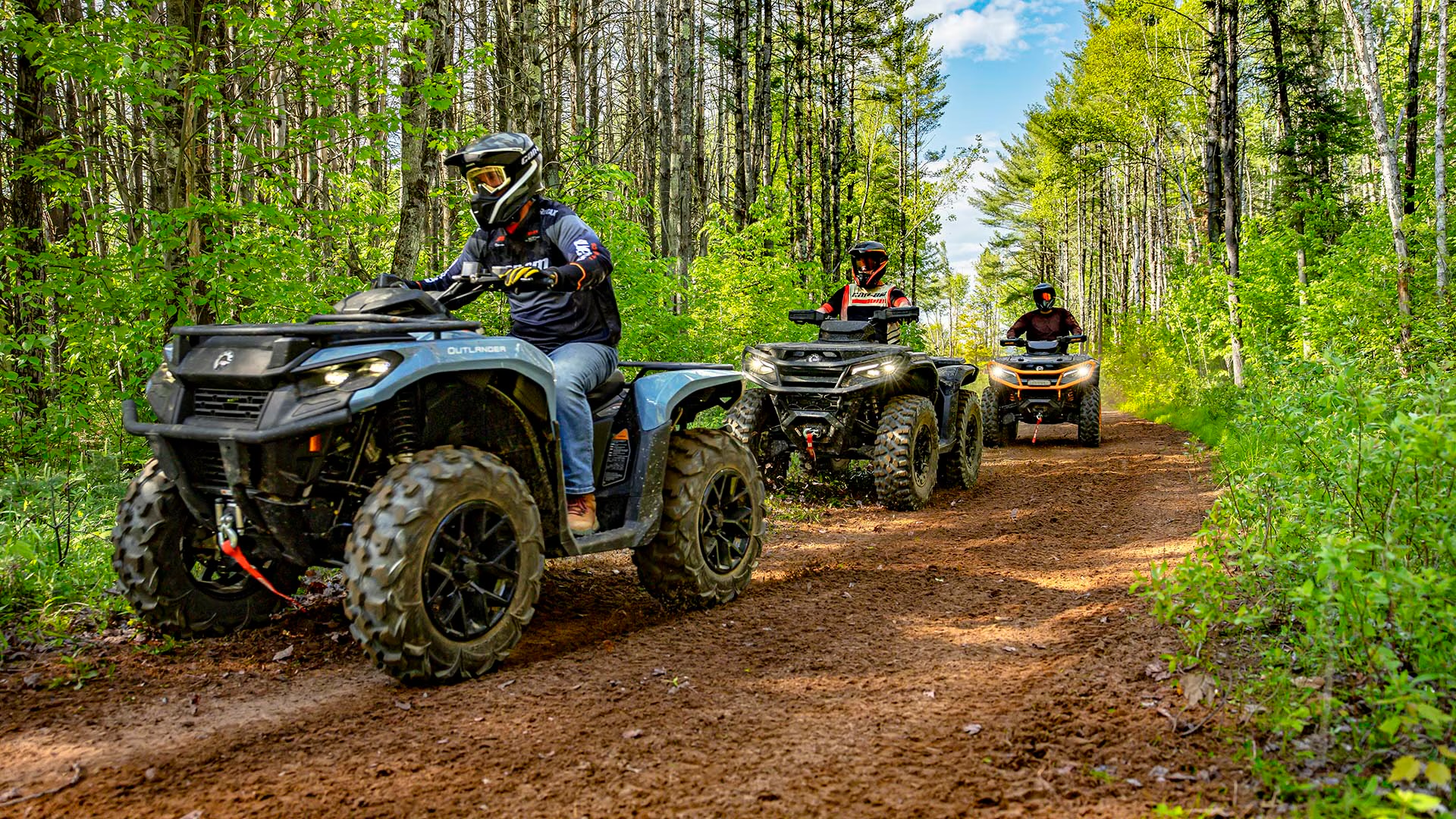 Três quadriciclos acelerando em fila na trilha de terra cercada de mata
